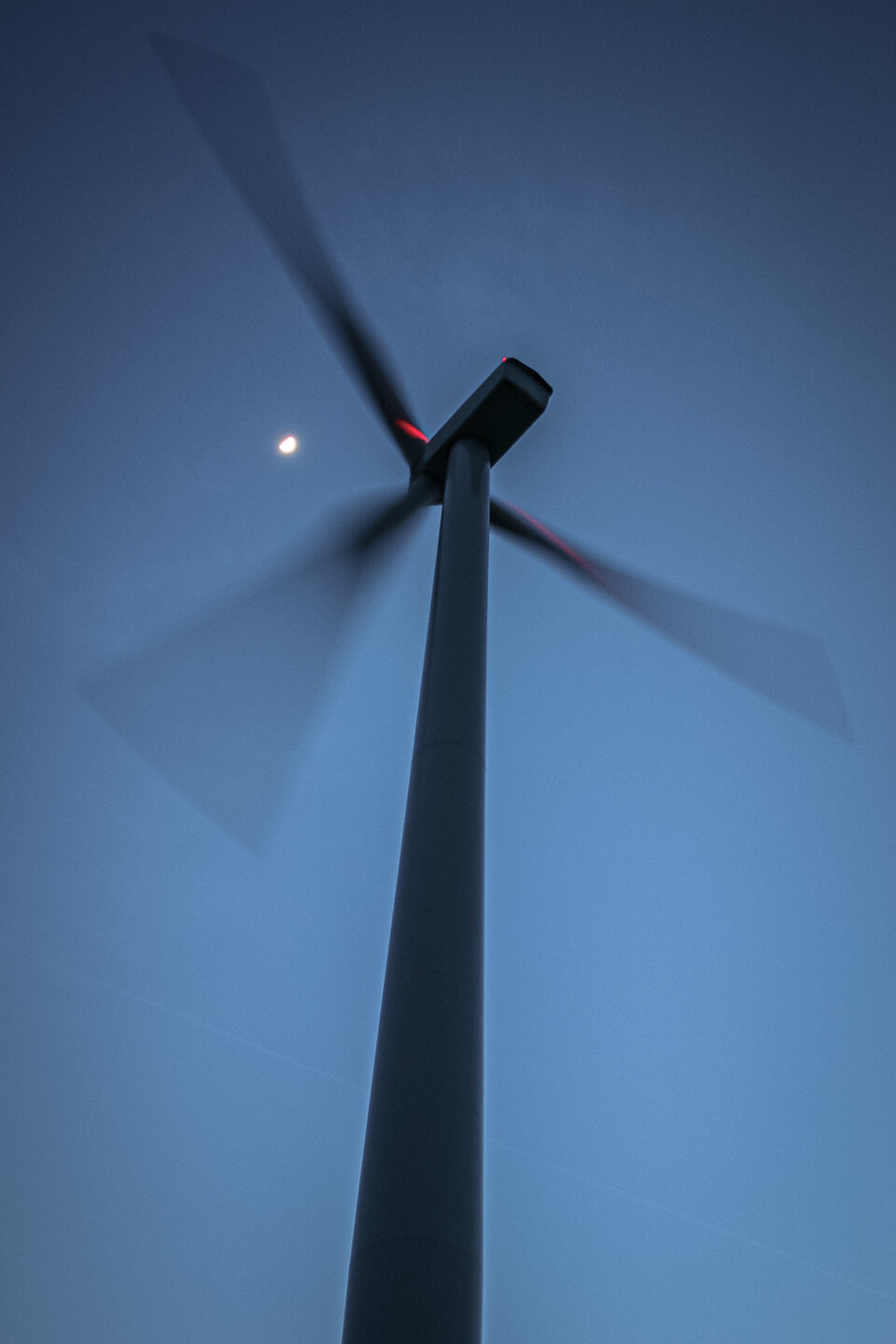 Contre-plongée d'une éolienne en mouvement à Sombreffe en Belgique, prise au crépuscule. Les pales floues indiquent le mouvement, avec la lune visible dans le ciel bleu foncé.