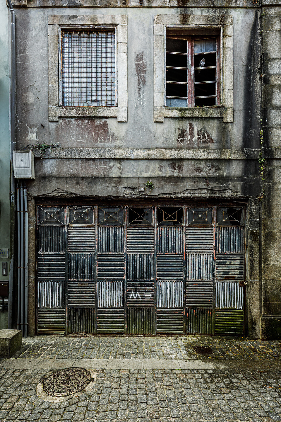 Façade usée en béton et pierre d'un bâtiment urbain au rez-de-chaussée fermé par une grande porte de garage pliante métallique à motifs géométriques striés, surmontée de deux fenêtres encadrées de pierre, dont une avec grillage et l'autre aux carreaux brisés où perche un pigeon, au-dessus d'une rue en pavés. (Vila Nove de Gaia, Porto, Portugal)