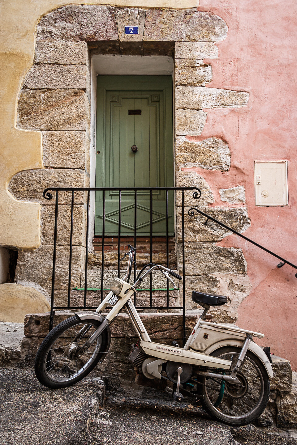 Une mobylette Peugeot 102 blanche garée devant une porte verte en bois avec le numéro 7, dans une rue pavée de Callas, dans le Var en France. Les murs sont en pierre et peints en rose et beige.