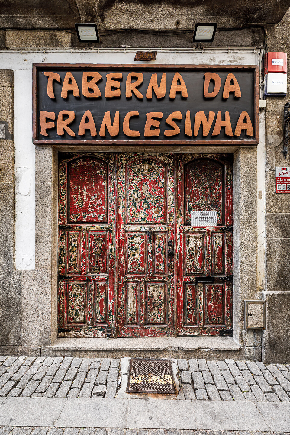 Vue de face au niveau de la rue d'une devanture de restaurant. En haut, une grande enseigne rectangulaire en bois présente le texte en relief « Taberna da Francesinha » en grosses lettres couleur bois clair sur un fond sombre. Au-dessous, une double porte en bois rouge et vieillie présente une texture poncée qui révèle de multiples couches de peintures colorées sous-jacentes d'époque. (Vila Nova de Gaia, Porto, Portugal)