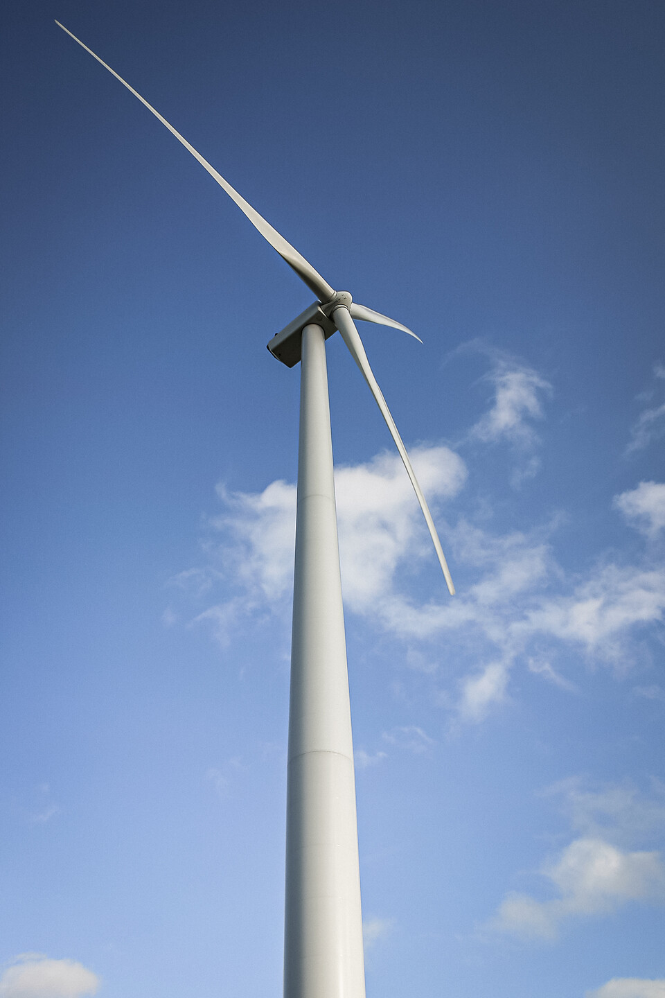 Contre-plongée d'une éolienne moderne à trois pales blanches à Sombreffe en Belgique, se détachant sur un ciel bleu clair avec quelques nuages blancs épars.