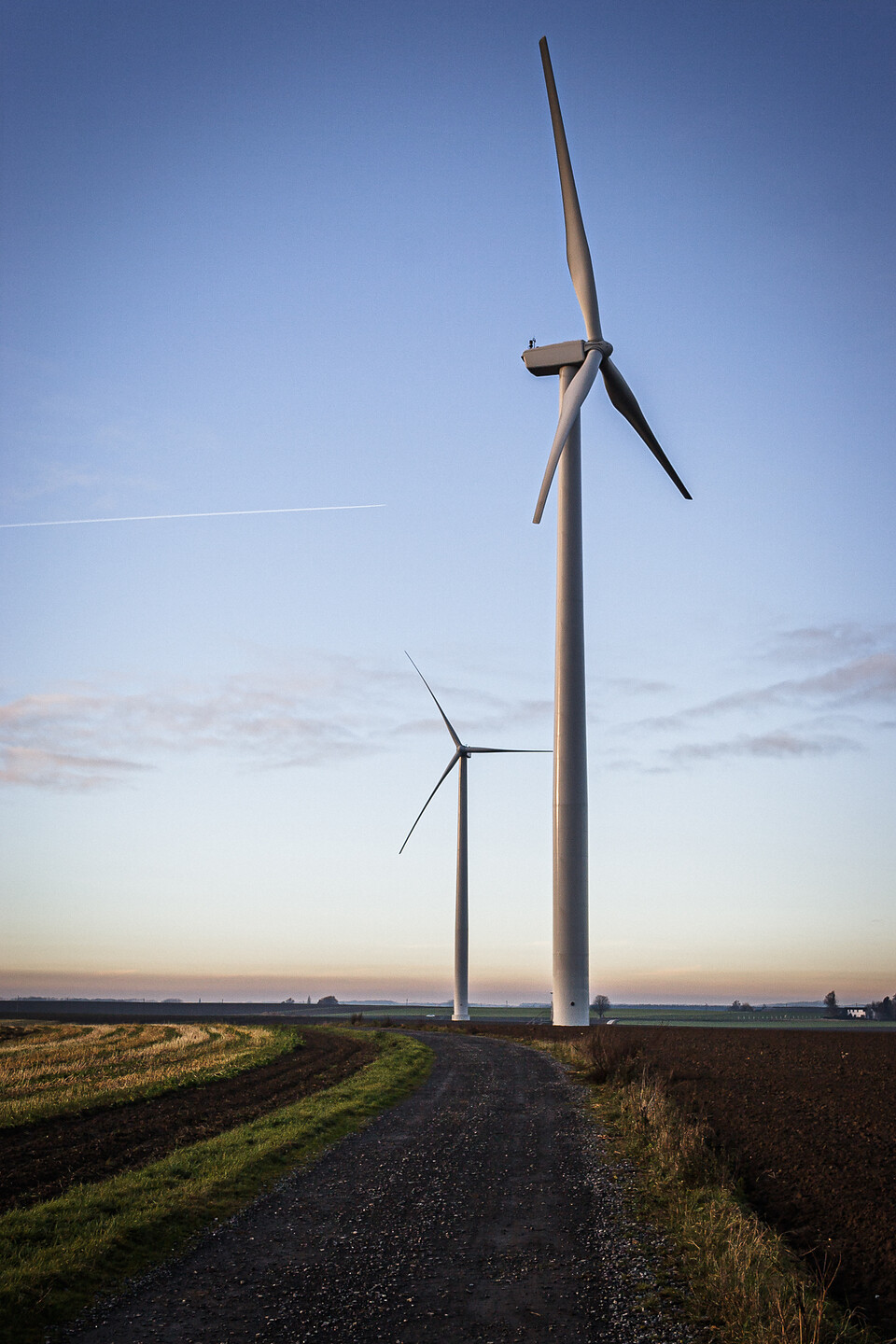 Paysage avec deux éoliennes modernes dans un champ à Sombreffe en Belgique, au crépuscule. Un chemin de gravier mène aux éoliennes, avec un ciel nuageux et des teintes chaudes du soleil couchant.
