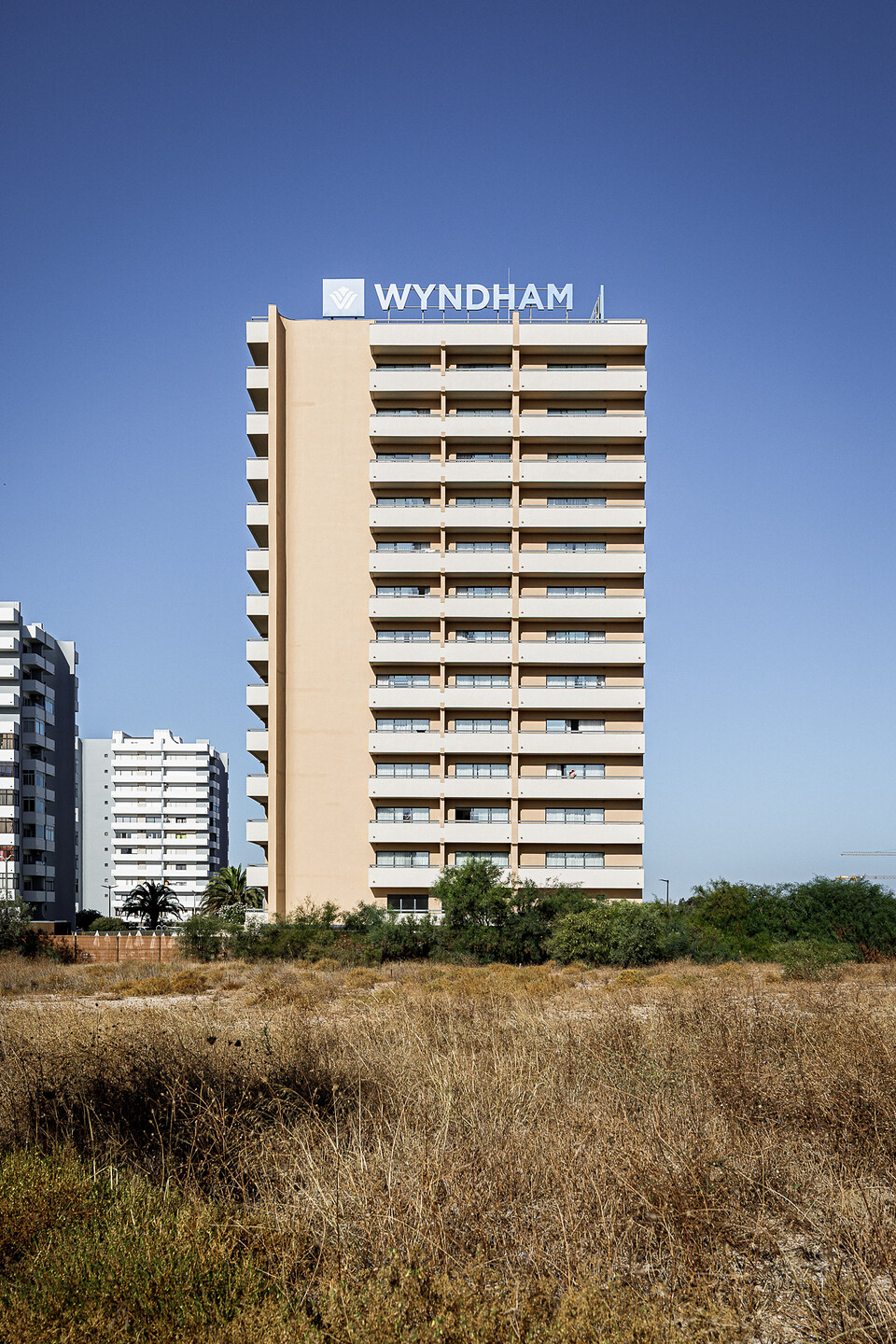 Façade principale d'un immeuble hôtelier Wyndham de plusieurs étages avec balcons alignés, sur fond de ciel bleu, derrière un terrain en friche aux hautes herbes sèches.