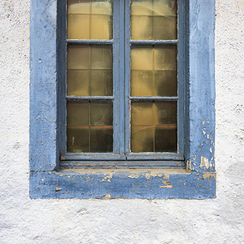 Plein cadre sur une fenêtre aux vitres jaunies et son encadrement peint en bleu sur un mur blanc.
