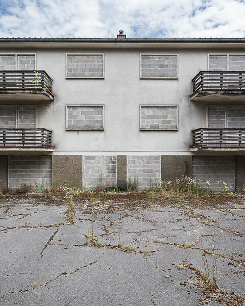 Vue d'ensemble symétrique du bâtiment abandonné au 30-32 route de Trèves. La structure de trois étages, murée à tous les niveaux, se dresse sous un ciel partiellement nuageux.