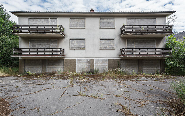 Vue d'ensemble symétrique du bâtiment abandonné au 30-32 route de Trèves. La structure de trois étages, murée à tous les niveaux, se dresse sous un ciel partiellement nuageux.