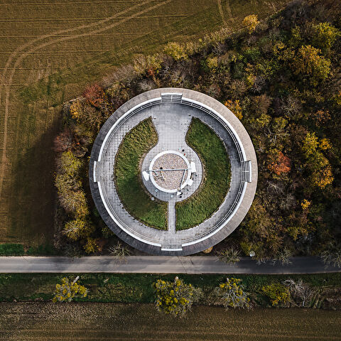 Vue aérienne du château d'eau de Remerschen, remarquable par sa plateforme au sommet composée de béton et pelouse. La structure est encadrée par une route, des champs labourés et une forêt aux couleurs d'automne.