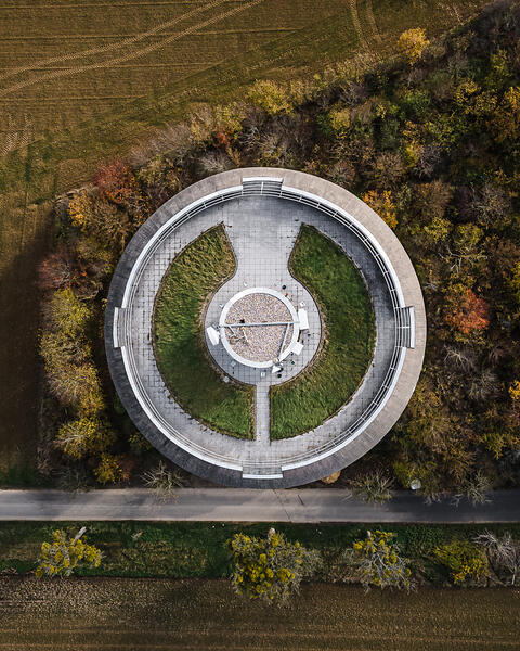 Vue aérienne du château d'eau de Remerschen, remarquable par sa plateforme au sommet composée de béton et pelouse. La structure est encadrée par une route, des champs labourés et une forêt aux couleurs d'automne.