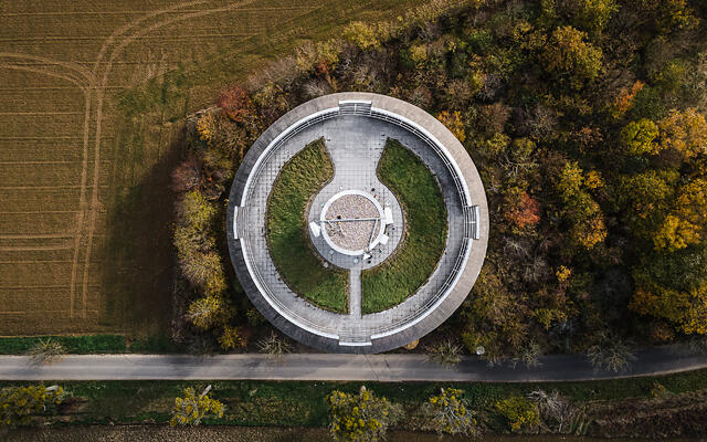 Vue aérienne du château d'eau de Remerschen, remarquable par sa plateforme au sommet composée de béton et pelouse. La structure est encadrée par une route, des champs labourés et une forêt aux couleurs d'automne.