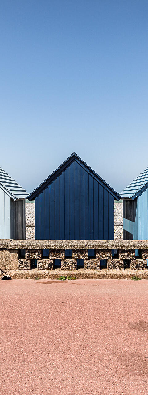 Une rangée de cabines de plage en bois aux dégradés de bleu, alignées derrière un muret en pierre sur le front de mer de Dieppe. Au centre, une cabine bleu marine se détache parmi d'autres aux tons bleu ciel et blanc cassé. Le premier plan montre une promenade rose saumoné sous un ciel bleu clair et dégagé.