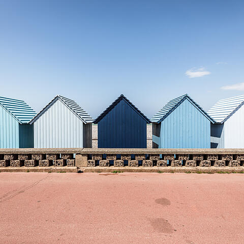 Une rangée de cabines de plage en bois aux dégradés de bleu, alignées derrière un muret en pierre sur le front de mer de Dieppe. Au centre, une cabine bleu marine se détache parmi d'autres aux tons bleu ciel et blanc cassé. Le premier plan montre une promenade rose saumoné sous un ciel bleu clair et dégagé.