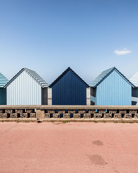 Une rangée de cabines de plage en bois aux dégradés de bleu, alignées derrière un muret en pierre sur le front de mer de Dieppe. Au centre, une cabine bleu marine se détache parmi d'autres aux tons bleu ciel et blanc cassé. Le premier plan montre une promenade rose saumoné sous un ciel bleu clair et dégagé.