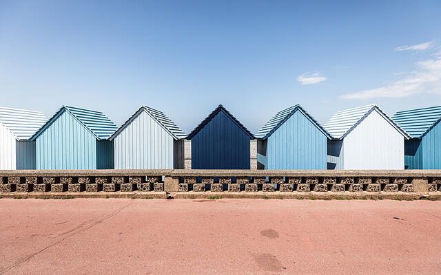 Une rangée de cabines de plage en bois aux dégradés de bleu, alignées derrière un muret en pierre sur le front de mer de Dieppe. Au centre, une cabine bleu marine se détache parmi d'autres aux tons bleu ciel et blanc cassé. Le premier plan montre une promenade rose saumoné sous un ciel bleu clair et dégagé.