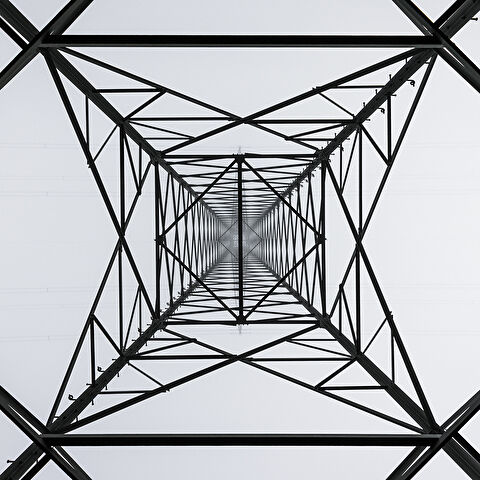 Vue en contre-plongée d'un pylône électrique, dont la structure métallique complexe forme une cage géométrique avec des lignes électriques horizontales.