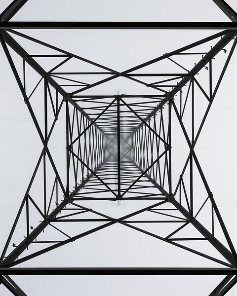 Vue en contre-plongée d'un pylône électrique, dont la structure métallique complexe forme une cage géométrique avec des lignes électriques horizontales.