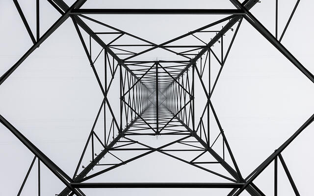 Vue en contre-plongée d'un pylône électrique, dont la structure métallique complexe forme une cage géométrique avec des lignes électriques horizontales.