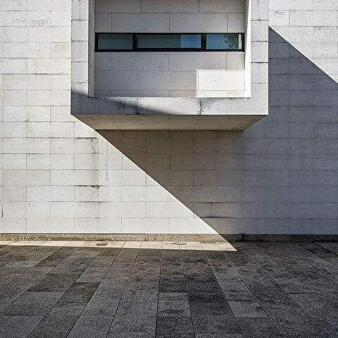 Façade en pierres calcaires blanches d'un bâtiment moderne (Centro de Memória de Vila do Conde, Porto, Portugal), avec un volume carré en saillie abritant une fenêtre horizontale. Une ombre triangulaire nette est projetée sous la saillie, accentuant le contraste avec le sol en dalles de granit foncé.