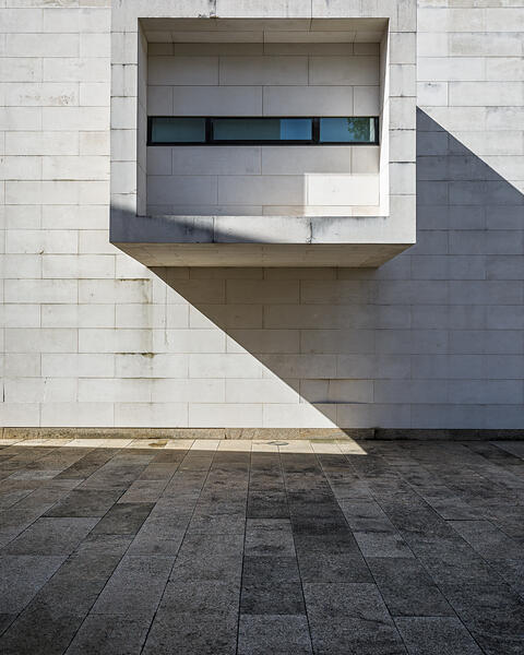 Façade en pierres calcaires blanches d'un bâtiment moderne (Centro de Memória de Vila do Conde, Porto, Portugal), avec un volume carré en saillie abritant une fenêtre horizontale. Une ombre triangulaire nette est projetée sous la saillie, accentuant le contraste avec le sol en dalles de granit foncé.