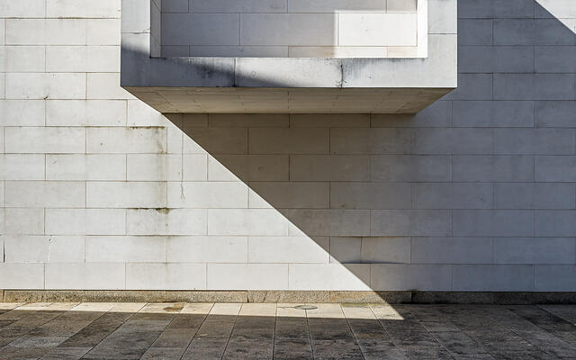 Façade en pierres calcaires blanches d'un bâtiment moderne (Centro de Memória de Vila do Conde, Porto, Portugal), avec un volume carré en saillie abritant une fenêtre horizontale. Une ombre triangulaire nette est projetée sous la saillie, accentuant le contraste avec le sol en dalles de granit foncé.