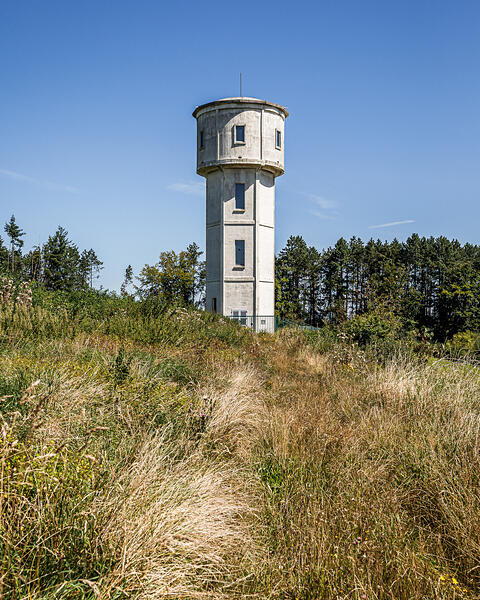 Château d'eau cylindrique en béton gris clair avec des fenêtres rectangulaires, situé au sommet d'une colline herbeuse jaune-brun à Frassem, Belgique, entouré d'une forêt de conifères et sous un ciel bleu clair.