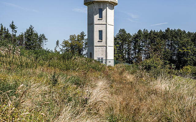Château d'eau cylindrique en béton gris clair avec des fenêtres rectangulaires, situé au sommet d'une colline herbeuse jaune-brun à Frassem, Belgique, entouré d'une forêt de conifères et sous un ciel bleu clair.