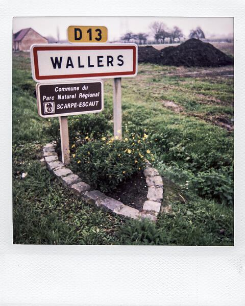 Polaroid montrant un panneau routier blanc bordé de rouge indiquant 'WALLERS', surmonté du cartouche jaune D13, situé dans un paysage rural.