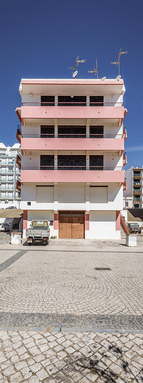 Immeuble résidentiel moderne de plusieurs étages aux façades blanches et roses, avec balcons, antennes paraboliques et véhicules stationnés devant, sur une place en calçada portuguesa.