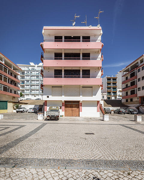 Immeuble résidentiel moderne de plusieurs étages aux façades blanches et roses, avec balcons, antennes paraboliques et véhicules stationnés devant, sur une place en calçada portuguesa.