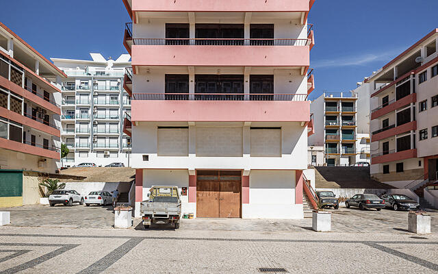 Façade symétrique d'un immeuble blanc et rose sur une place en pavés, surmonté d'antennes télé sous un ciel bleu dégagé. (Monte Gordo, Portugal)