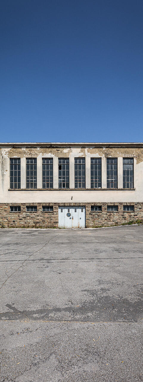 Vue frontale du bâtiment technique abandonné de l'ancienne laiterie Laduno-Luxlait, avec sa façade en pierre et en crépi défraîchi, ses grandes fenêtres verticales et sa cour asphaltée sous un ciel bleu.