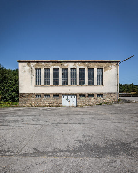 Vue frontale du bâtiment technique abandonné de l'ancienne laiterie Laduno-Luxlait, avec sa façade en pierre et en crépi défraîchi, ses grandes fenêtres verticales et sa cour asphaltée sous un ciel bleu.