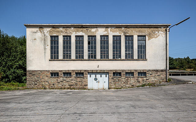 Ancienne laiterie Laduno-Luxlait. Façade d'un bâtiment industriel abandonné avec soubassement en pierre, grandes fenêtres et porte métallique sur un parking en asphalte. (Erpeldange-sur-Sure, Luxembourg)