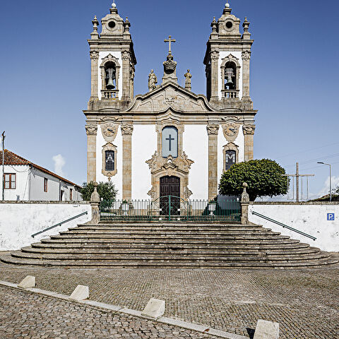 Église baroque avec deux clochers, façade ornée de statues, entrée centrale en bois surmontée d’une croix, située au sommet d’escaliers en pierre. Cour pavée, ciel bleu, arbres et bâtiment à gauche, panneau de stationnement à droite.