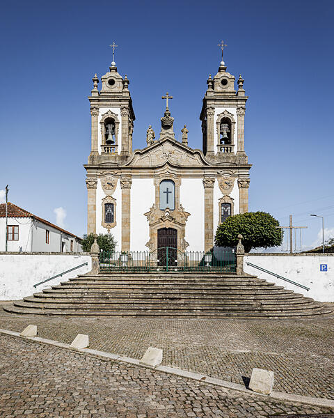 Église baroque avec deux clochers, façade ornée de statues, entrée centrale en bois surmontée d’une croix, située au sommet d’escaliers en pierre. Cour pavée, ciel bleu, arbres et bâtiment à gauche, panneau de stationnement à droite.