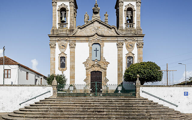 Église baroque avec deux clochers, façade ornée de statues, entrée centrale en bois surmontée d'une croix, située au sommet d'escaliers en pierre. Cour pavée, ciel bleu, arbres et bâtiment à gauche, panneau de stationnement à droite.