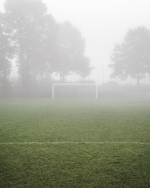 La photo représente un terrain de football par temps de brouillard. Le gazon est vert, un but blanc se trouve au centre et des arbres en arrière-plan sont partiellement cachés par le brouillard.