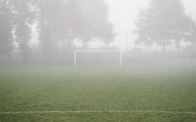 La photo représente un terrain de football par temps de brouillard. Le gazon est vert, un but blanc se trouve au centre et des arbres en arrière-plan sont partiellement cachés par le brouillard.
