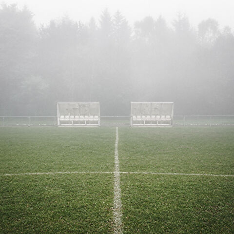 L'image montre un terrain de football embrumé, avec deux bancs d'équipe vides placés de part et d'autre de la ligne médiane. Le terrain est recouvert d'herbe verte, et une ligne blanche marquant le centre divise l'image verticalement. À l'arrière-plan, une forêt dense d'arbres hauts est partiellement masquée par le brouillard.