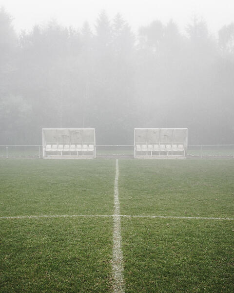L'image montre un terrain de football embrumé, avec deux bancs d'équipe vides placés de part et d'autre de la ligne médiane. Le terrain est recouvert d'herbe verte, et une ligne blanche marquant le centre divise l'image verticalement. À l'arrière-plan, une forêt dense d'arbres hauts est partiellement masquée par le brouillard.