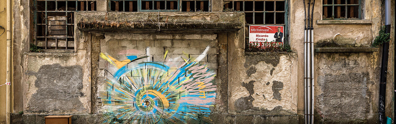 Ancienne façade délabrée à Vila Nova de Gaia, près de Porto, avec murs en pierre et enduit écaillé, fenêtres à petits carreaux aux vitres brisées, traces d’humidité et de végétation. Un graffiti coloré bleu, jaune et rose orne une ouverture murée au centre du bâtiment, tandis qu’une affiche de l’agence immobilière MéritoInvest apparaît dans une fenêtre à droite. Des rubans de sécurité barrent le trottoir au premier plan.