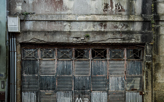 Façade usée en béton et pierre d'un bâtiment urbain au rez-de-chaussée fermé par une grande porte de garage pliante métallique à motifs géométriques striés, surmontée de deux fenêtres encadrées de pierre, dont une avec grillage et l'autre aux carreaux brisés où perche un pigeon, au-dessus d'une rue en pavés. (Vila Nova de Gaia, Porto, Portugal)