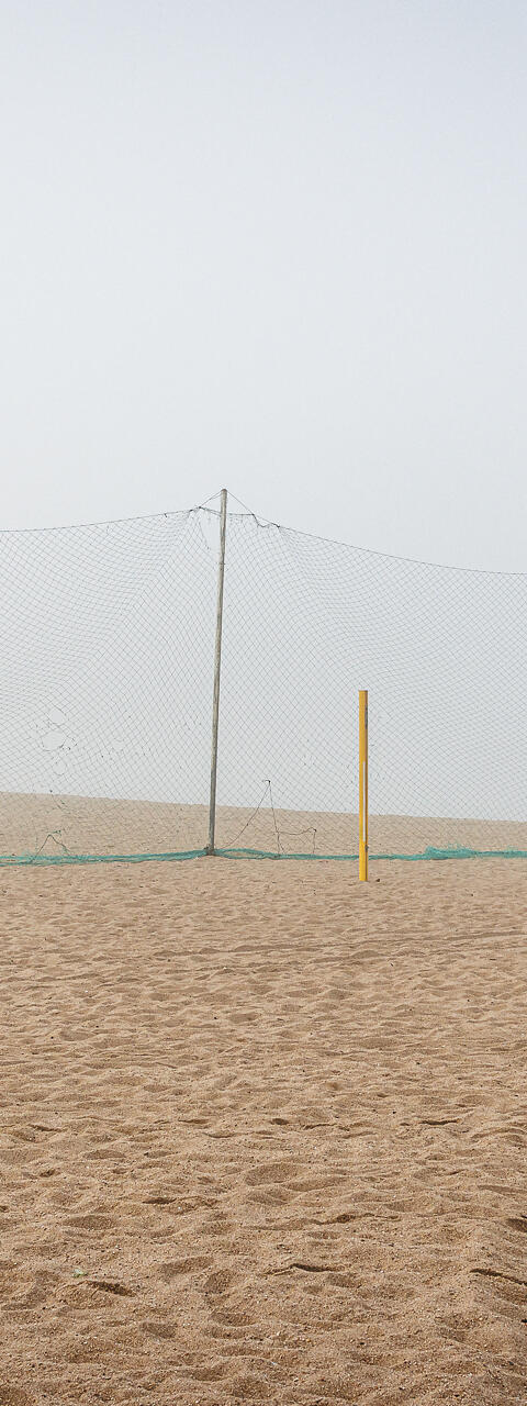 Plan large et minimaliste d'un terrain de beach-volley déserté, sous une lumière diffuse due au brouillard. Au centre, un filet vert est tendu pour séparer le terrain de la plage. Deux poteaux de couleur jaune, sans filet, se dressent à droite dans le sable.