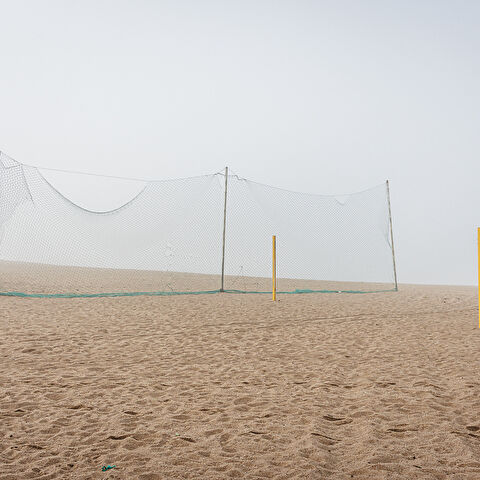 Plan large et minimaliste d'un terrain de beach-volley déserté, sous une lumière diffuse due au brouillard. Au centre, un filet vert est tendu pour séparer le terrain de la plage. Deux poteaux de couleur jaune, sans filet, se dressent à droite dans le sable.