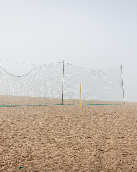 Plan large et minimaliste d'un terrain de beach-volley déserté, sous une lumière diffuse due au brouillard. Au centre, un filet vert est tendu pour séparer le terrain de la plage. Deux poteaux de couleur jaune, sans filet, se dressent à droite dans le sable.