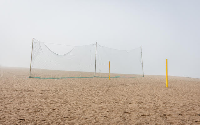 Plan large et minimaliste d'un terrain de beach-volley déserté, sous une lumière diffuse due au brouillard. Au centre, un filet vert est tendu pour séparer le terrain de la plage. Deux poteaux de couleur jaune, sans filet, se dressent à droite dans le sable.