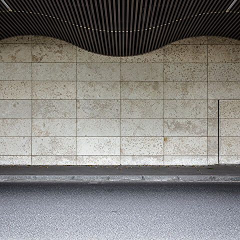 un pan de mur composé de carreaux rectangulaires avec un motif tacheté. Une petite porte est visible sur le côté droit du mur. Au-dessus du mur, on distingue une structure ondulée et sombre faite de lattes, qui fait partie du toit. Le sol est pavé d'asphalte, et une bordure sépare le mur de la route.