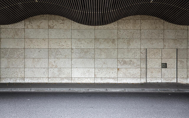 Un pan de mur composé de carreaux rectangulaires avec un motif tacheté. Une petite porte est visible sur le côté droit du mur. Au-dessus du mur, on distingue une structure ondulée et sombre faite de lattes, qui fait partie du toit. Le sol est pavé d'asphalte, et une bordure sépare le mur de la route.