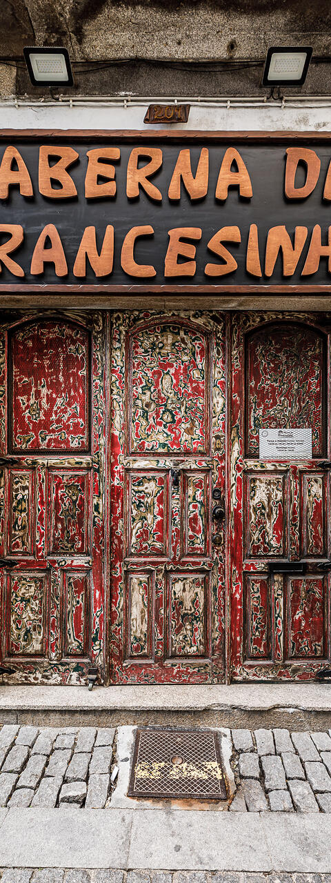 Vue de face au niveau de la rue d'une devanture de restaurant. En haut, une grande enseigne rectangulaire en bois présente le texte en relief « Taberna da Francesinha » en grosses lettres couleur bois clair sur un fond sombre. Au-dessous, une double porte en bois rouge et vieillie présente une texture poncée qui révèle de multiples couches de peintures colorées sous-jacentes d'époque. (Vila Nova de Gaia, Porto, Portugal)