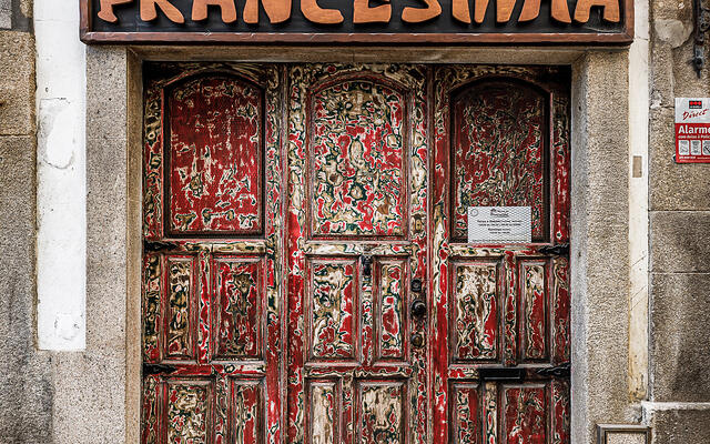 Vue de face au niveau de la rue d'une devanture de restaurant. En haut, une grande enseigne rectangulaire en bois présente le texte en relief « Taberna da Francesinha » en grosses lettres couleur bois clair sur un fond sombre. Au-dessous, une double porte en bois rouge et vieillie présente une texture poncée qui révèle de multiples couches de peintures colorées sous-jacentes d'époque. (Vila Nova de Gaia, Porto, Portugal)