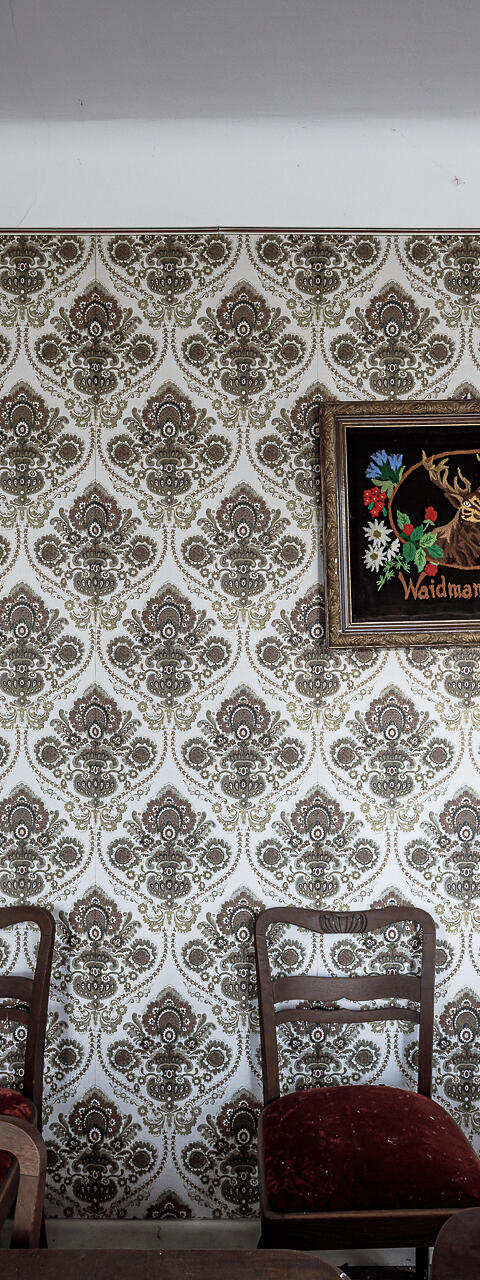 Austère salle à manger meublée de chaises aux coussins de velours rouge. Avec pour seule décoration un cadre illustré 'Waidmanns Heil!' ...